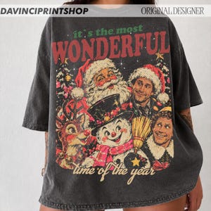 K&ouml;nnte beinhalten: Ein anthrazitfarbenes Oversize-T-Shirt mit einem Grafikdruck von Weihnachtsmann, Rentier, Schneemann und Buddy dem Elf. Der Text lautet "it's the most WONDERFUL time of the year" in Rot und Gr&uuml;n. In den Ecken befinden sich Textbeschriftungen.