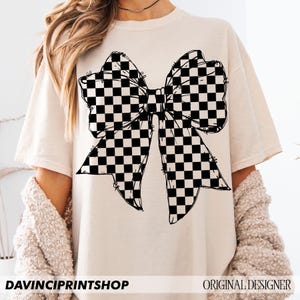 May include: Cream-colored t-shirt featuring a large black and white checkered bow graphic. The shirt has short sleeves and a relaxed fit. The text "DAVINCIPRINTSHOP" is at the bottom left, and "ORIGINAL DESIGNER" is at the bottom right.