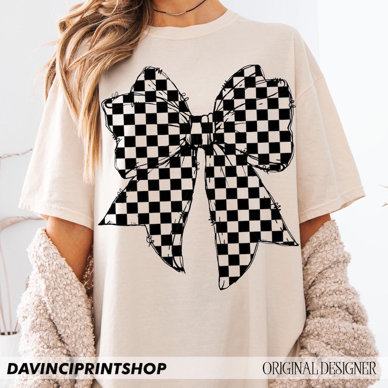 May include: Cream-colored t-shirt featuring a large black and white checkered bow graphic. The shirt has short sleeves and a relaxed fit. The text "DAVINCIPRINTSHOP" is at the bottom left, and "ORIGINAL DESIGNER" is at the bottom right.
