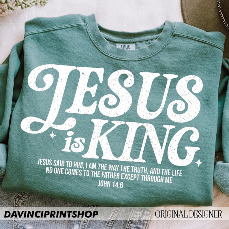 K&ouml;nnte beinhalten: Ein t&uuml;rkisfarbenes Sweatshirt mit einer wei&szlig;en Grafik, auf der in einer Retro-Schrift "JESUS is KING" steht. Die Grafik enth&auml;lt auch einen Bibelvers: "Jesus sagte zu ihm: Ich bin der Weg, die Wahrheit und das Leben. Niemand kommt zum Vater au&szlig;er durch mich. Johannes 14:6"