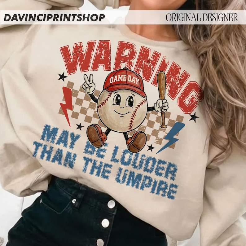 K&ouml;nnte beinhalten: Cremefarbenes Sweatshirt mit Baseball-Motiv. Die Grafik zeigt eine Baseball-Figur mit roter Kappe und Baseballschl&auml;ger. Der Text enth&auml;lt "WARNING" und "MAY BE LOUDER THAN THE UMPIRE".