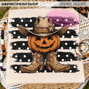 K&ouml;nnte beinhalten: Ein beiges T-Shirt mit einer Grafik eines K&uuml;rbisses, der einen braunen Cowboyhut und braune Cowboystiefel tr&auml;gt. Das Design ist vor einem schwarzen horizontalen Pinselstrich-Hintergrund mit wei&szlig;en Punkten platziert. Der Text lautet DAVINCIPRINTSHOP.
