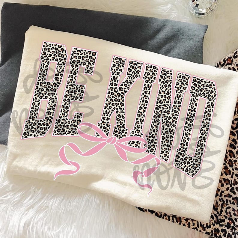 Op de afbeelding: Cr&egrave;mekleurig T-shirt met de tekst "BE KIND" in een luipaardprint ontwerp omlijnd in roze. Een roze strik is gecentreerd onder de tekst. Het shirt is op een grijze ondergrond gevouwen.
