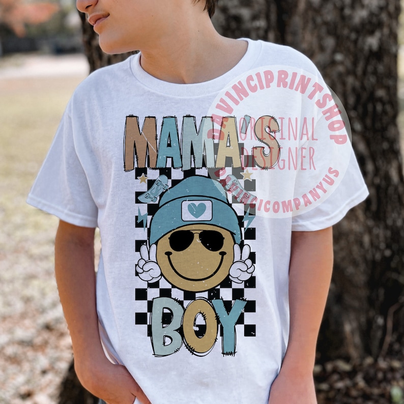 May include: White t-shirt with a graphic design featuring a smiling face wearing sunglasses and a blue beanie. The graphic also includes the text "Mama's Boy" in colourful letters and a checkered pattern.