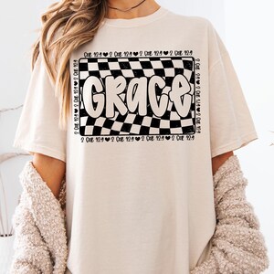 Op de afbeelding: Een wit T-shirt met een zwart-wit geruit ontwerp en het woord "Grace" in een zwart, bubbelvormig lettertype. Het geruite ontwerp is omgeven door de tekst "2 Cor 12:9" die meerdere keren wordt herhaald.