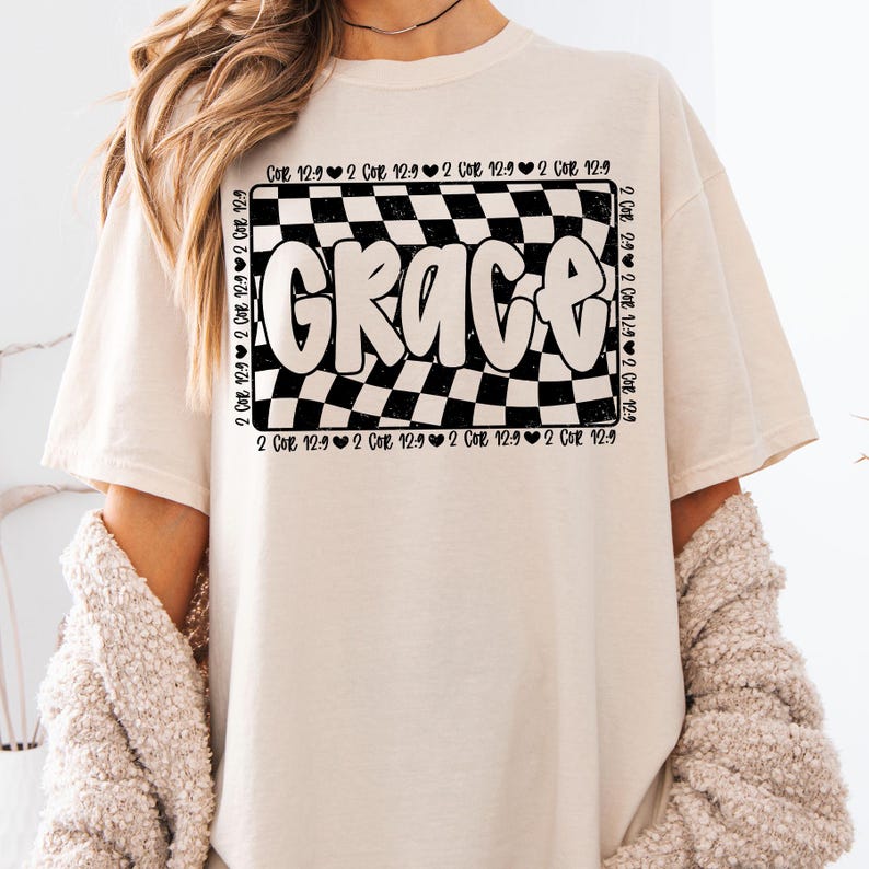 Op de afbeelding: Een wit T-shirt met een zwart-wit geruit ontwerp en het woord "Grace" in een zwart, bubbelvormig lettertype. Het geruite ontwerp is omgeven door de tekst "2 Cor 12:9" die meerdere keren wordt herhaald.