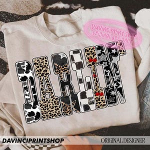 May include: A beige sweatshirt with the word "DARLIN" in a bold, patterned design. The letters feature cow print, leopard print, stars, and checkered patterns. The text "DAVINCIPRINTSHOP ORIGINAL DESIGNER" is also visible.