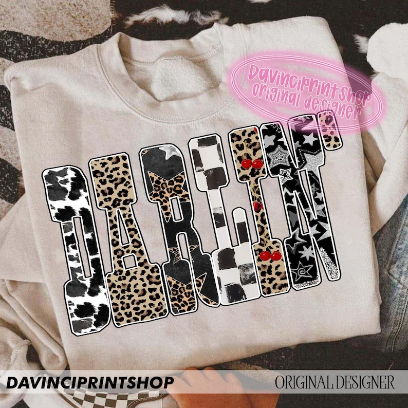 May include: A beige sweatshirt with the word "DARLIN" in a bold, patterned design. The letters feature cow print, leopard print, stars, and checkered patterns. The text "DAVINCIPRINTSHOP ORIGINAL DESIGNER" is also visible.