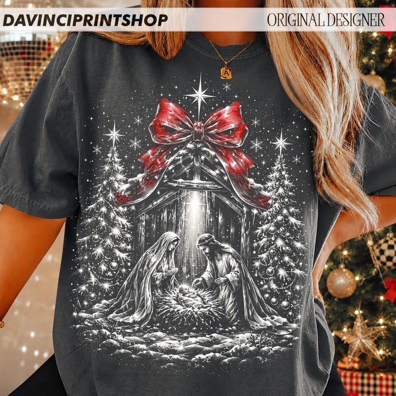 May include: A charcoal gray t-shirt featuring a graphic print of a nativity scene with two figures kneeling over a manger, flanked by two Christmas trees. A large red bow sits above the scene, with white stars and light rays throughout the design.