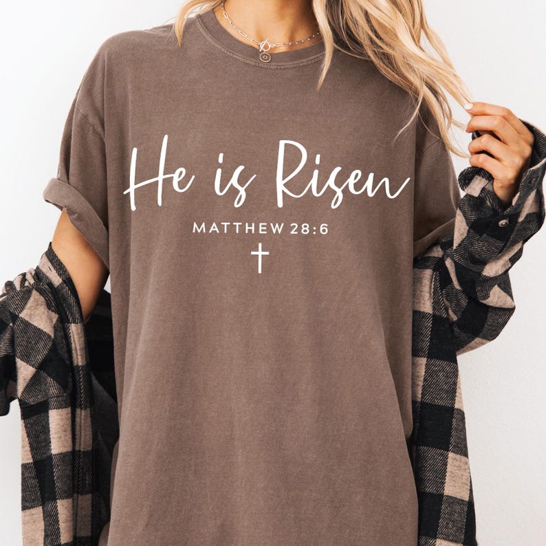 May include: A brown t-shirt with the text "He is Risen" and "Matthew 28:6" in white lettering. A small white cross is below the text.