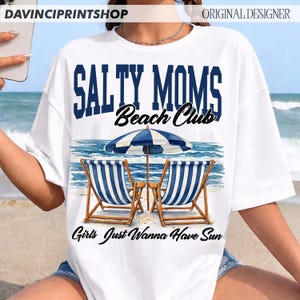 Op de afbeelding: Wit T-shirt met de tekst "SALTY MOMS Beach Club" in marineblauw en zwart. Het shirt heeft een strandsc&egrave;ne met twee blauw-wit gestreepte strandstoelen, een blauw-witte parasol en de zin "Girls Just Wanna Have Sun."