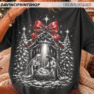 May include: A charcoal gray t-shirt featuring a graphic print of a nativity scene with Mary and Joseph surrounding a baby in a manger. The scene is framed by two Christmas trees, a large red bow at the top, and falling snowflakes.