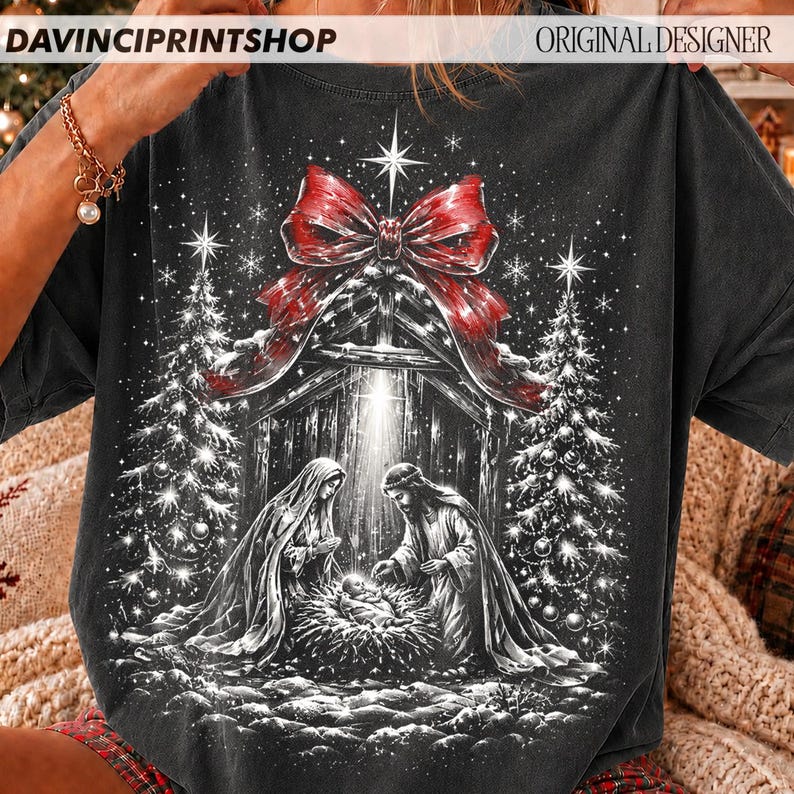 May include: A charcoal gray t-shirt featuring a graphic print of a nativity scene with Mary and Joseph surrounding a baby in a manger. The scene is framed by two Christmas trees, a large red bow at the top, and falling snowflakes.