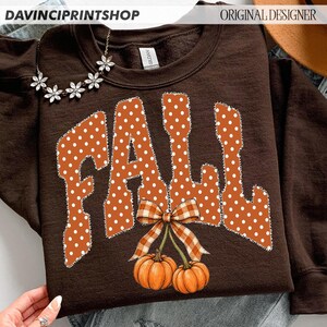 May include: A dark brown crewneck sweatshirt featuring the word FALL in large orange letters with white polka dots. Below the text are two orange pumpkins with green stems and a brown and white checkered bow tied around the stems.