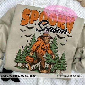 May include: Beige sweatshirt with the words "SPOOKY SEASON" in orange and black. A Bigfoot-like creature wearing a witch hat and pumpkin hoodie walks through a forest, carrying a jack-o'-lantern. Bats and stars are also featured.