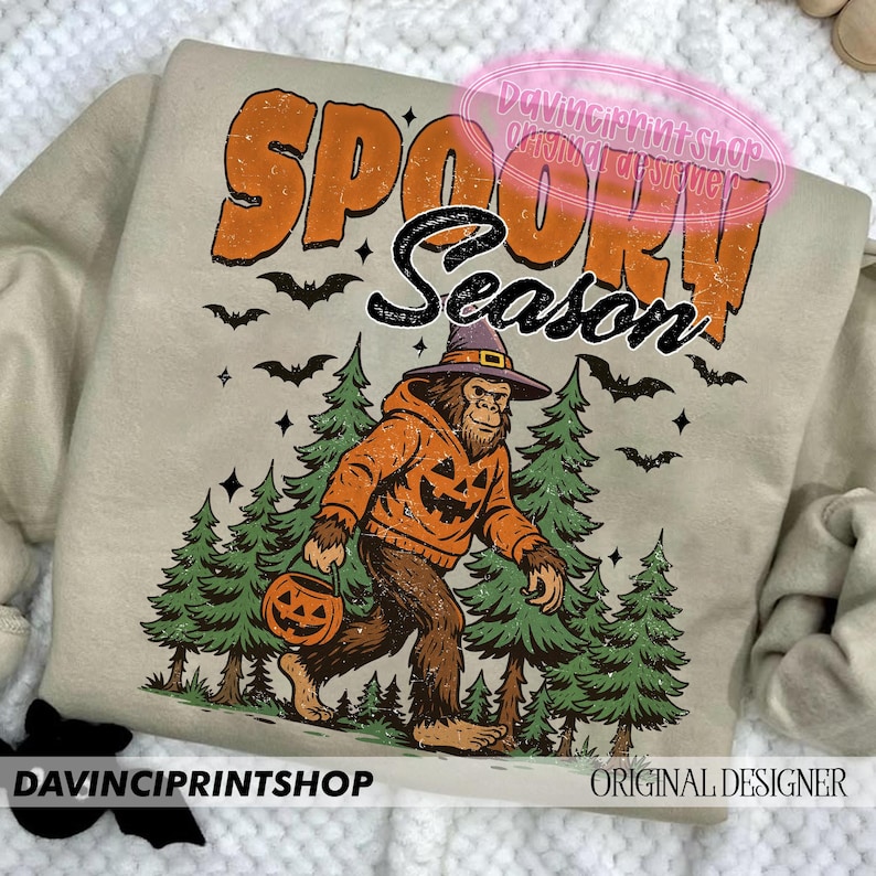 May include: Beige sweatshirt with the words "SPOOKY SEASON" in orange and black. A Bigfoot-like creature wearing a witch hat and pumpkin hoodie walks through a forest, carrying a jack-o'-lantern. Bats and stars are also featured.