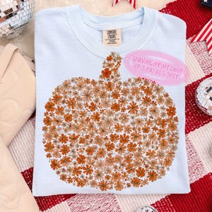 May include: Light blue Comfort Colors sweatshirt featuring a pumpkin design made of orange and peach-colored flowers. The text "DaVinciPrintShop Original Designer" is on the upper right. The shirt is on a red and white checkered surface.