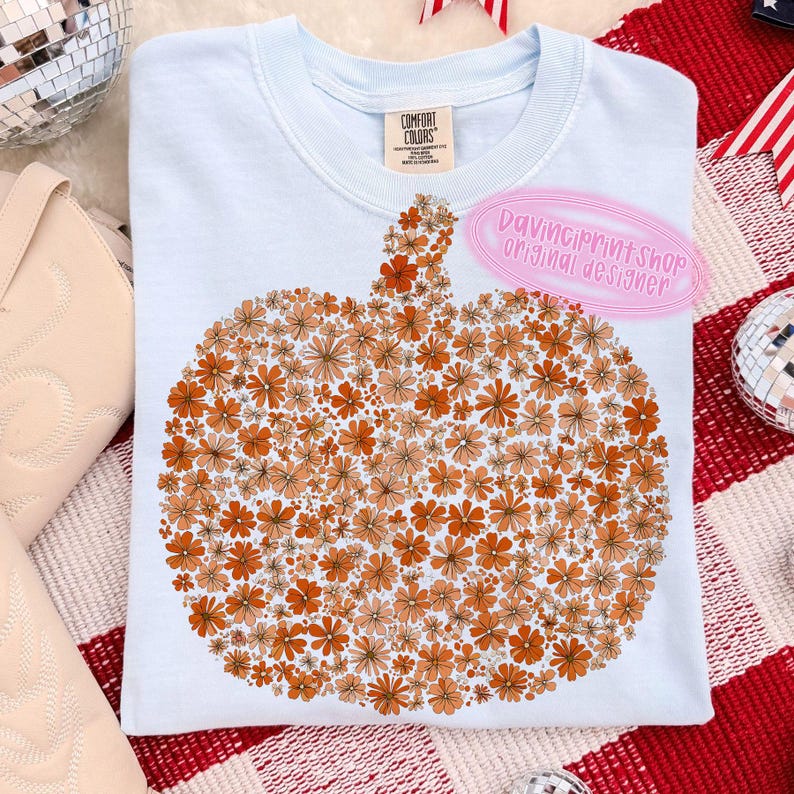 May include: Light blue Comfort Colors sweatshirt featuring a pumpkin design made of orange and peach-colored flowers. The text "DaVinciPrintShop Original Designer" is on the upper right. The shirt is on a red and white checkered surface.