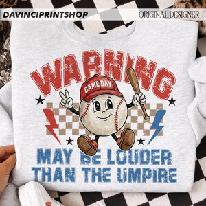 K&ouml;nnte beinhalten: Ein hellgraues Sweatshirt mit einem Baseball-Motiv. Das Motiv zeigt einen Baseball-Charakter mit roter Kappe und einem Schl&auml;ger. Der Text lautet "WARNING" und "MAY BE LOUDER THAN THE UMPIRE".