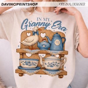Puede incluir: Camiseta beige con una ilustraci&oacute;n caprichosa. El dise&ntilde;o presenta un pato con un lazo azul, una estanter&iacute;a con tazas y teteras, y el texto "IN MY Granny Era" en azul. El tema general es vintage y acogedor.