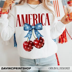 May include: White sweatshirt with the word "AMERICA" in red letters and a blue and white bow with stars above two red cherries with white stars.