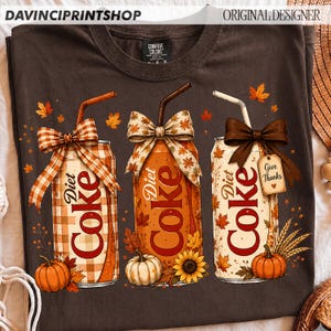 May include: A dark brown t-shirt featuring a graphic of three soda cans decorated with autumn themes. Each can has a straw, a bow, and fall elements like pumpkins, sunflowers, and leaves. One can has a tag that reads Give Thanks.