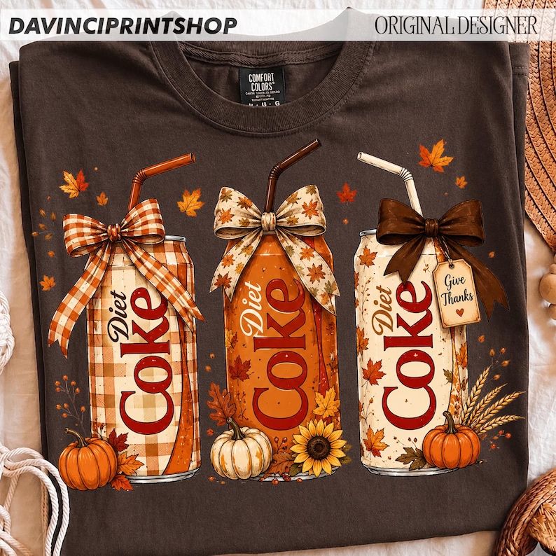 May include: A dark brown t-shirt featuring a graphic of three soda cans decorated with autumn themes. Each can has a straw, a bow, and fall elements like pumpkins, sunflowers, and leaves. One can has a tag that reads Give Thanks.