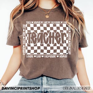 May include: A brown t-shirt with a white checkered design and the word "Teacher" in a white, bubbly font. The checkered design is surrounded by a white circle with the words "Teach, Inspire, Care, Encourage, Lead, Support, Teach, Care, Listen, Support, Teach, Care, Listen, Lead, Encourage, Inspire" written around it.