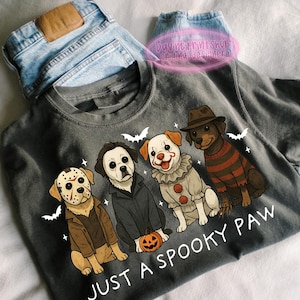 May include: Dark gray t-shirt featuring four cartoon dogs dressed as horror movie characters. The dogs are dressed as Jason Voorhees, Michael Myers, Pennywise the Clown, and Freddy Krueger. The text "JUST A SPOOKY PAW" is printed below the dogs.