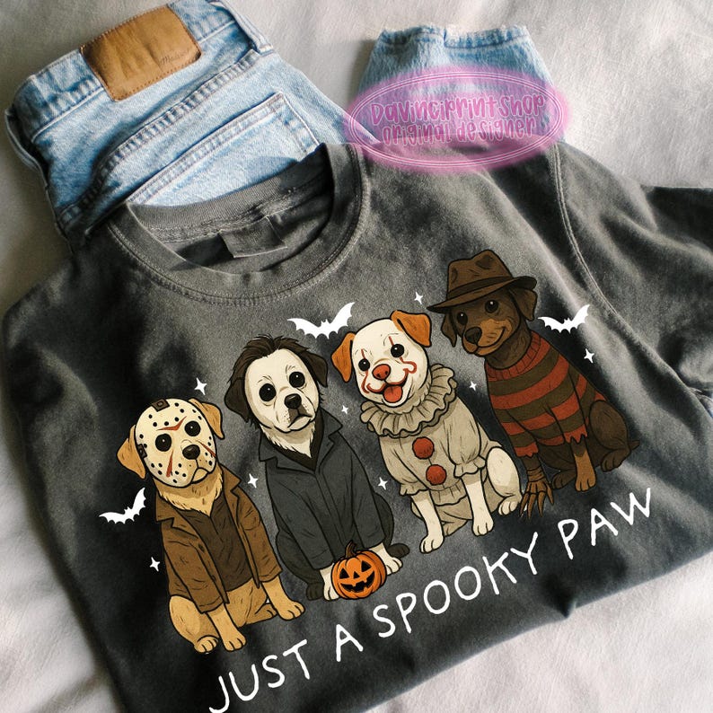 May include: Dark gray t-shirt featuring four cartoon dogs dressed as horror movie characters. The dogs are dressed as Jason Voorhees, Michael Myers, Pennywise the Clown, and Freddy Krueger. The text "JUST A SPOOKY PAW" is printed below the dogs.