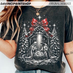 May include: A charcoal gray t-shirt featuring a graphic print of a nativity scene with a large red bow at the top. The design includes two figures kneeling over a manger, flanked by two decorated Christmas trees under a bright star.