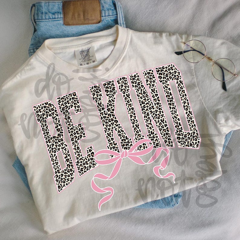 Op de afbeelding: Cr&egrave;mekleurig T-shirt met de tekst "BE KIND" in een luipaardprint ontwerp, omlijnd in roze. Een roze strik staat onder de tekst. Het shirt ligt op een lichtblauwe jeans.