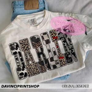 May include: Cream-colored t-shirt with the word "DARE" in a bold, block letter design. Each letter features a unique pattern, including leopard print, cow print, and checkered patterns. The shirt is laid on top of blue jeans.