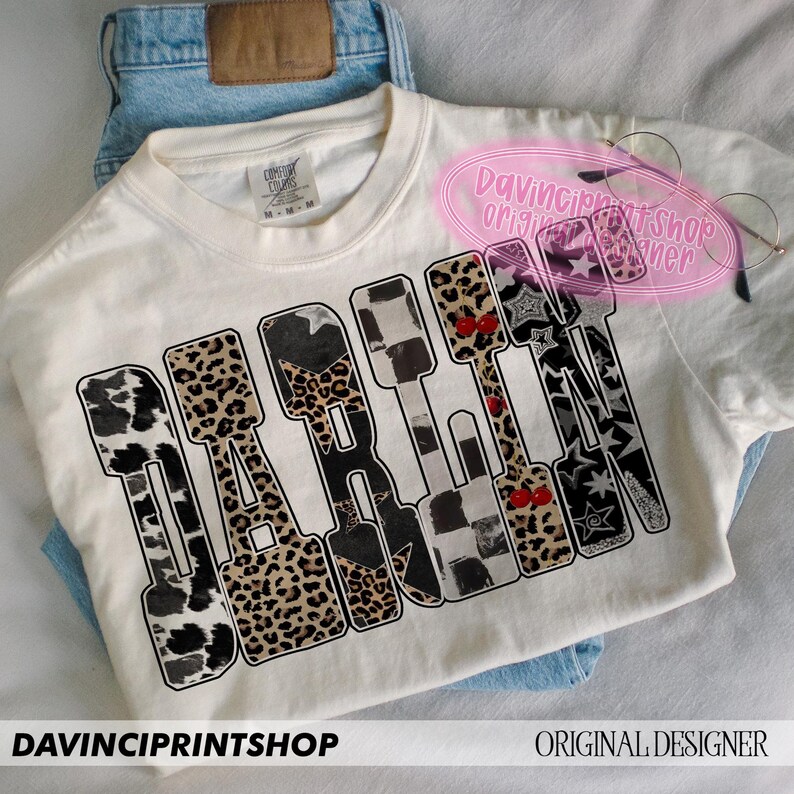 May include: Cream-colored t-shirt with the word "DARE" in a bold, block letter design. Each letter features a unique pattern, including leopard print, cow print, and checkered patterns. The shirt is laid on top of blue jeans.