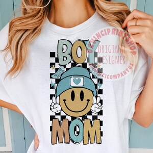 May include: White t-shirt featuring a checkered background, a smiling face wearing a blue beanie with a heart on it, and the words "Boy Mom" in colourful letters.