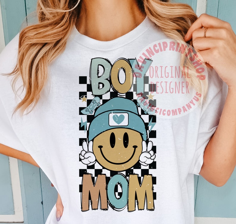 May include: White t-shirt featuring a checkered background, a smiling face wearing a blue beanie with a heart on it, and the words "Boy Mom" in colourful letters.