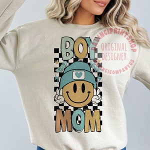 May include: A light beige sweatshirt featuring a graphic design of a smiling face wearing a blue beanie with a heart on it. The graphic is surrounded by a black and white chequered pattern and the words "Boy Mom" are printed below the face.