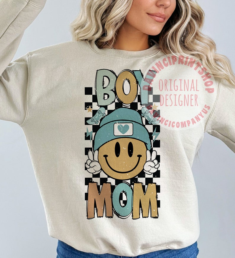 May include: A light beige sweatshirt featuring a graphic design of a smiling face wearing a blue beanie with a heart on it. The graphic is surrounded by a black and white chequered pattern and the words "Boy Mom" are printed below the face.