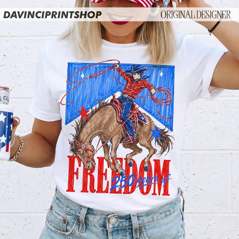 May include: White t-shirt featuring a graphic of a cowboy riding a horse, roping. The design includes red, white, and blue colors, with the word "FREEDOM" in large red letters and "250 years" below. The shirt is a casual wear item.