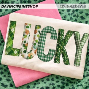 May include: A cream-colored shirt with the word "LUCKY" appliqued in various green patterns, including gingham and floral designs. The shirt is resting on a pink surface, with the text "DAVINCIPRINTSHOP" and "ORIGINAL DESIGNER" at the top.