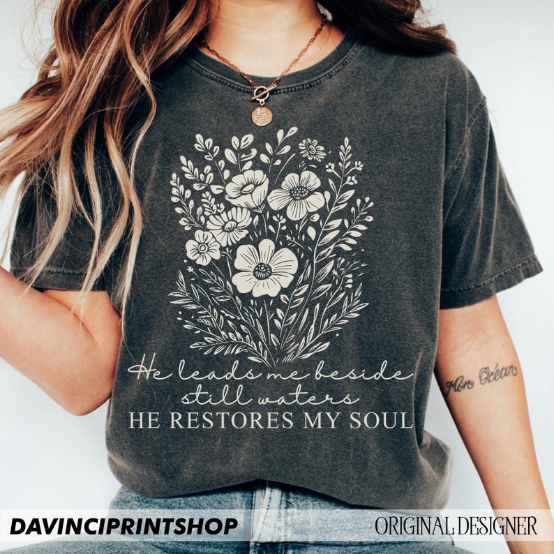 May include: A dark gray t-shirt with a floral design and the text "He leads me beside still waters HE RESTORES MY SOUL".