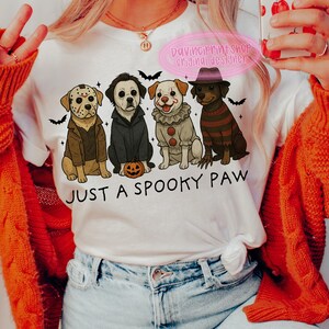 May include: White t-shirt with a Halloween-themed graphic. The design features four dogs dressed as horror movie characters, with bats and a pumpkin. The text "JUST A SPOOKY PAW" is printed below the dogs.