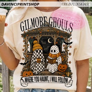Puede incluir: Una camiseta color crema con un gr&aacute;fico de dos fantasmas en un cenador con el texto "GILMORE GHOULS" y "WHERE YOU HAUNT, I WILL FOLLOW". El dise&ntilde;o incluye calabazas, hojas de oto&ntilde;o, murci&eacute;lagos, una luna creciente y un farol.