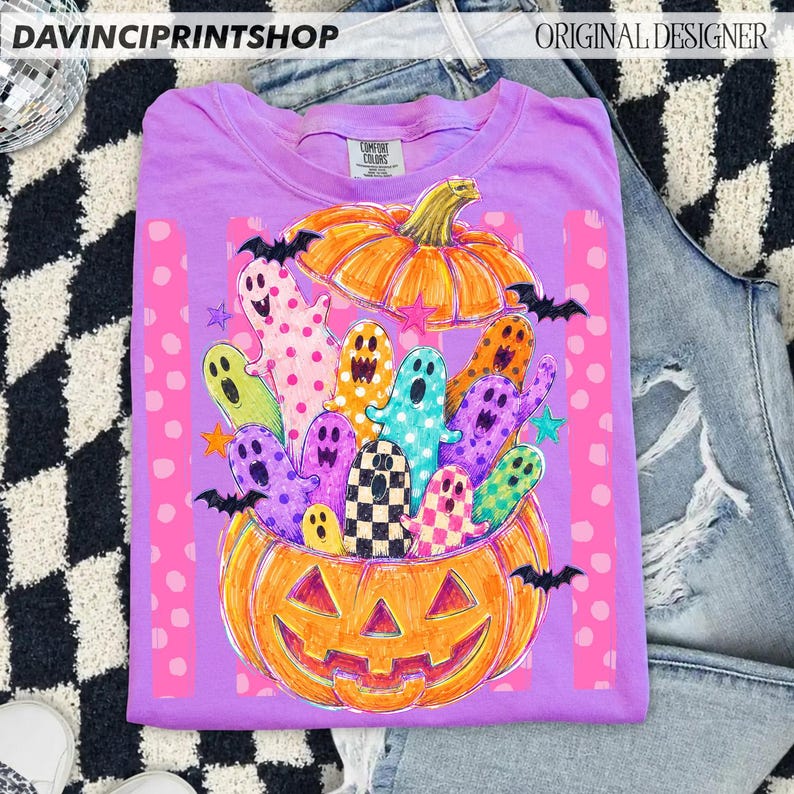 May include: A light purple t-shirt featuring a graphic of a large orange jack-o'-lantern filled with colorful, patterned ghosts and bats. The design includes pink vertical stripes with polka dots and small stars, creating a festive Halloween scene.