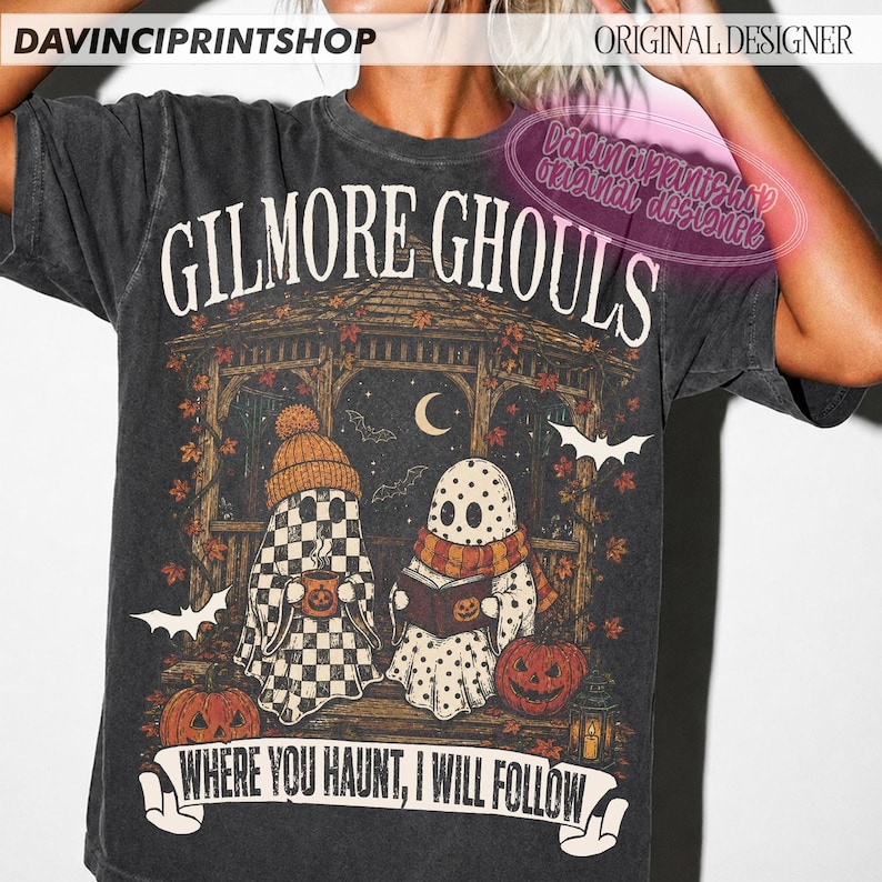 Puede incluir: Una camiseta gris carb&oacute;n con un estampado gr&aacute;fico que incluye el texto "GILMORE GHOULS" y "WHERE YOU HAUNT, I WILL FOLLOW". El dise&ntilde;o muestra dos fantasmas en un cenador con hojas de oto&ntilde;o, calabazas, murci&eacute;lagos y una luna creciente.