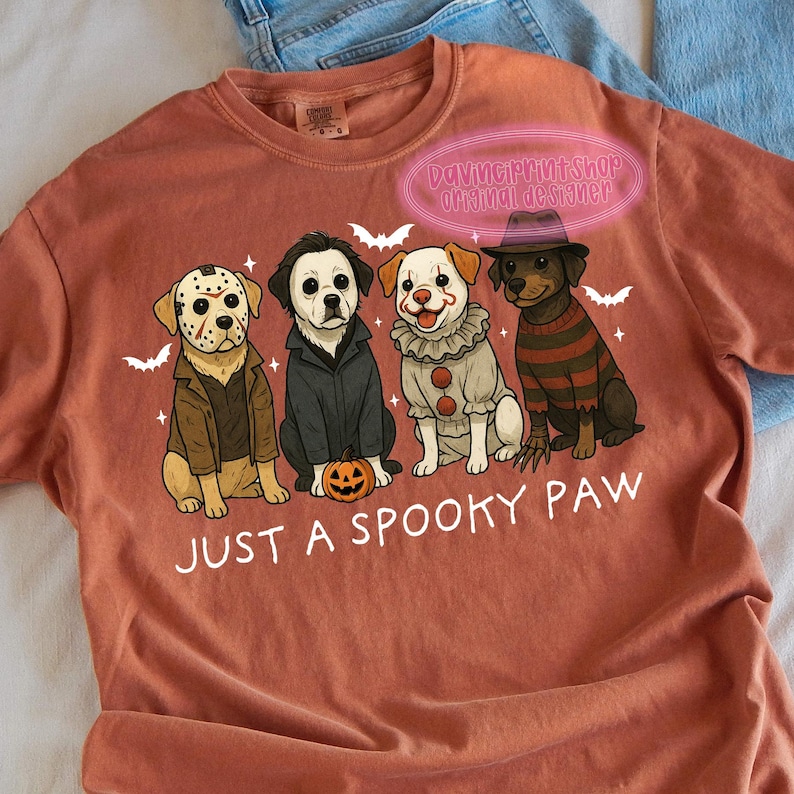 May include: A rust-colored t-shirt featuring a graphic of four dogs dressed as horror movie characters. The phrase "JUST A SPOOKY PAW" is printed below the dogs. Bats are also included in the design.