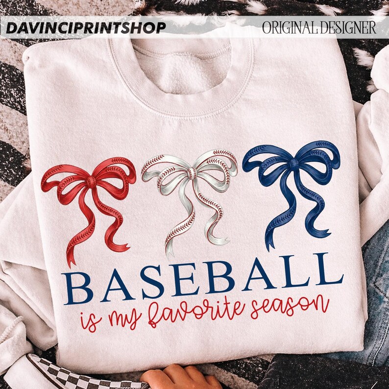 May include: White sweatshirt featuring three decorative bows in red, white (baseball design), and blue. The text "BASEBALL is my favorite season" is printed below the bows in navy and red script. The sweatshirt has a crew neck.
