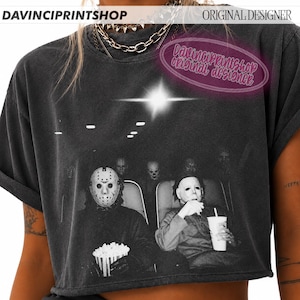 Peut inclure: Un t-shirt court gris anthracite avec une impression graphique de deux personnages masqu&eacute;s assis dans un cin&eacute;ma, l'un portant un masque de hockey avec du pop-corn, l'autre un masque blanc avec une boisson, sous une lumi&egrave;re vive.