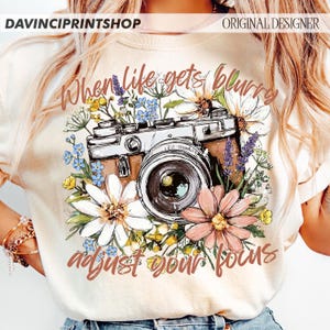 May include: Cream-colored t-shirt featuring a vintage camera design surrounded by colorful flowers. The text "When life gets blurry, adjust your focus" is printed in a stylish font. The shirt is a photography-themed apparel item.
