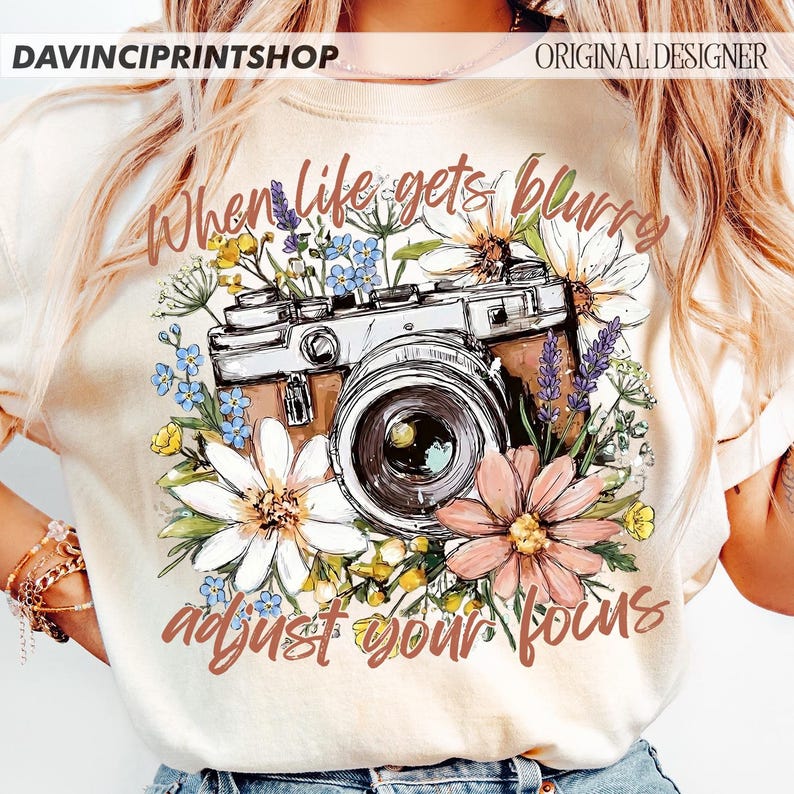 May include: Cream-colored t-shirt featuring a vintage camera design surrounded by colorful flowers. The text "When life gets blurry, adjust your focus" is printed in a stylish font. The shirt is a photography-themed apparel item.